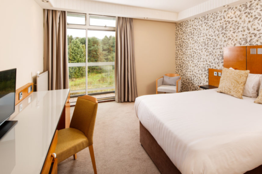Classic Double Room at Mercure Leeds Parkway Hotel, grey armchair beside bed, white desk and floor length windows with views onto garden