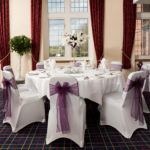 Table set up for wedding breakfast, white with purple sashes and floral centrepiece, next to window overlooking hills